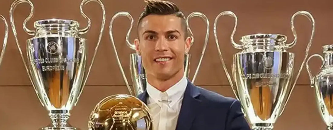 Soccer player in suit holding Ballon d'Or trophy, standing with UEFA Champions League cups