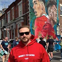 Man wearing a red hoodie with text in front of a mural of Bobby Firmino on a brick building.